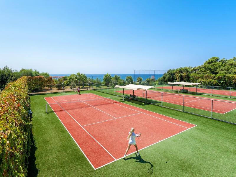Mehrere rote Tennisplätze mit grüner Einfassung und Blick auf das blaue Meer bei klarem Himmel.
