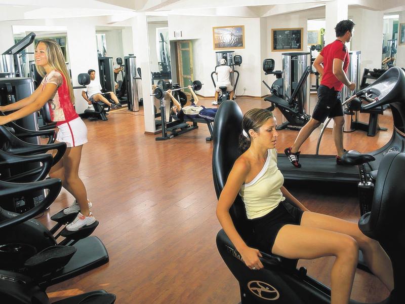 Fitnessraum mit Menschen auf Laufbändern und Trainingsgeräten, heller Raum mit Holzfußboden.