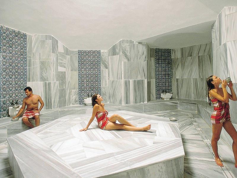 Innenraum eines türkischen Hamams mit weiß-grauen Marmorsäulen, Boden und Wänden, drei Personen in Badesachen.