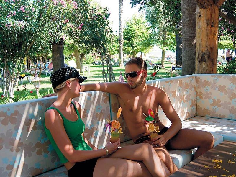 Zwei Personen sitzen sommerlich bekleidet auf einer hellen Outdoor-Lounge mit Bäumen im Hintergrund.