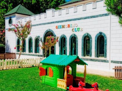 Área de juegos infantil con casita verde frente a un edificio blanco con ventanas arqueadas.