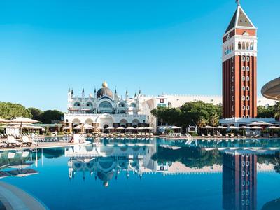 Area piscina con lettini di fronte a un hotel con torre sotto un cielo blu terso