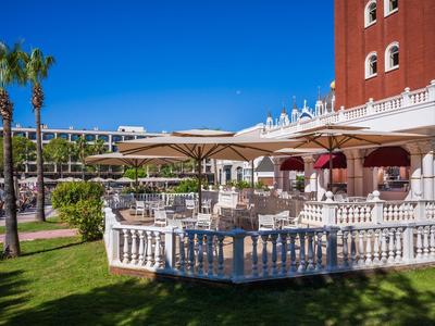 Słoneczny taras z miejscami do siedzenia przed czerwonym budynkiem hotelu i zielonymi palmami.