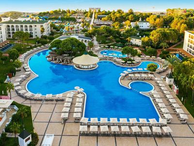 Duży niebieski basen z parasolami i leżakami w hotelowym ośrodku otoczonym drzewami.