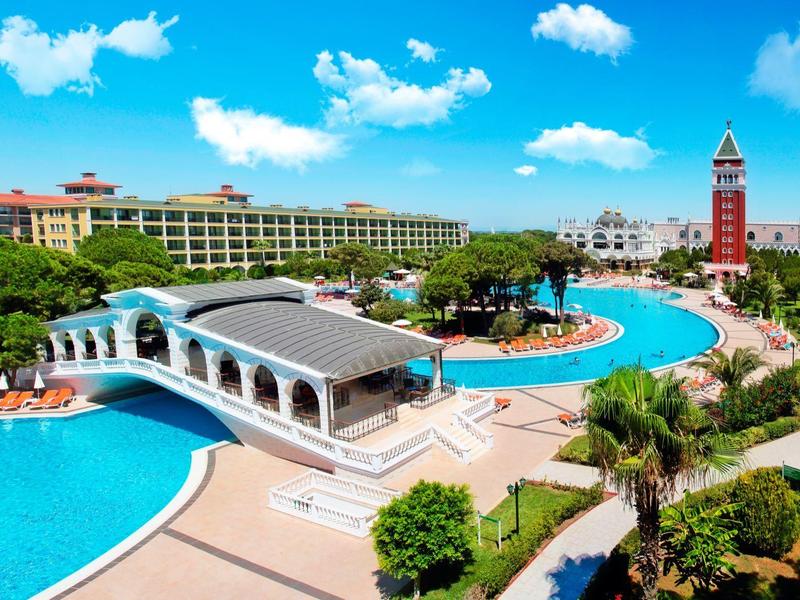 Gran piscina de hotel con puente, sombrillas y torre contigua bajo un cielo azul.