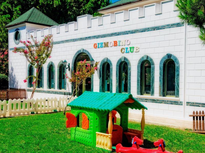 Área de juegos infantil con casita verde frente a un edificio blanco con ventanas arqueadas.