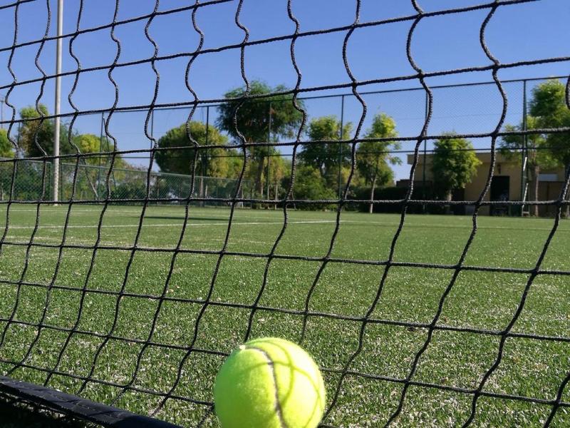 Pelota amarilla de tenis en la base de una portería de fútbol sobre el campo verde.