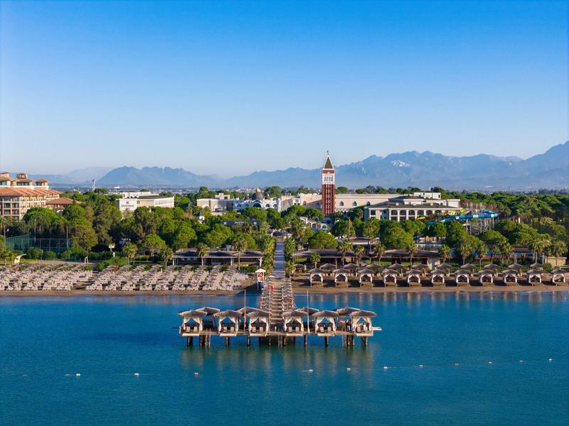 Vista de una zona costera con un muelle y tumbonas frente a un hotel y montañas al fondo.
