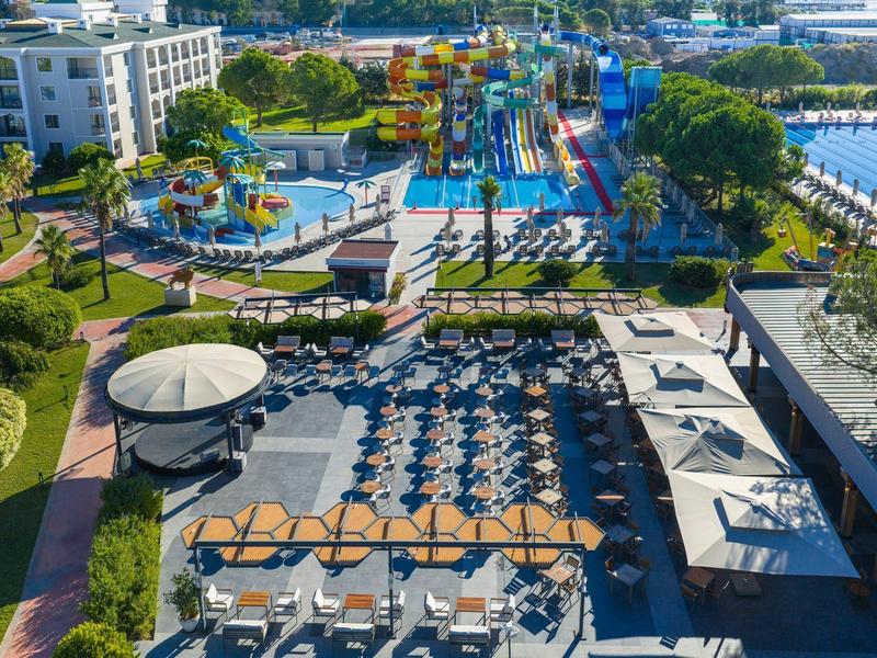 Vista aérea del área de piscina de un hotel con tumbonas, sombrillas y parque acuático al fondo.