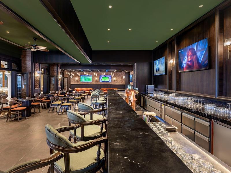 Zona de bar moderna con barra larga, sillas cómodas y pantallas LED en las paredes.