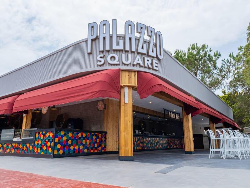 Entrada de un hotel o restaurante con gran letrero 'Palazzo Square' y toldos rojos.