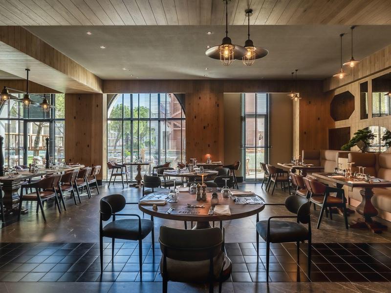 Restaurante elegante con mesas de madera, sillas y grandes ventanas que dejan entrar la luz del día.