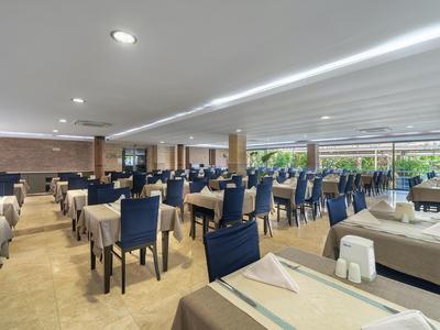 Großer heller Speisesaal mit blauen Stühlen, braunen Tischdecken und großen Fenstern.