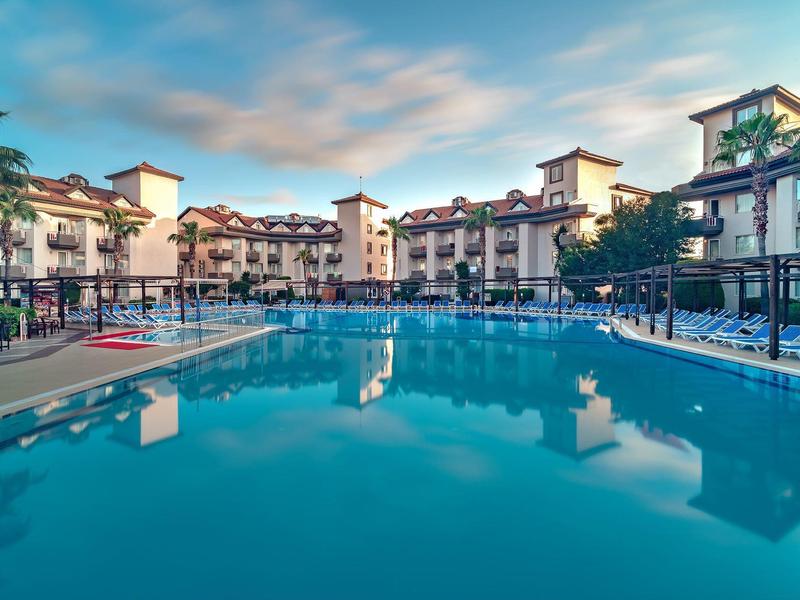 Gran piscina de hotel con tumbonas alrededor y edificios de varios pisos al fondo.