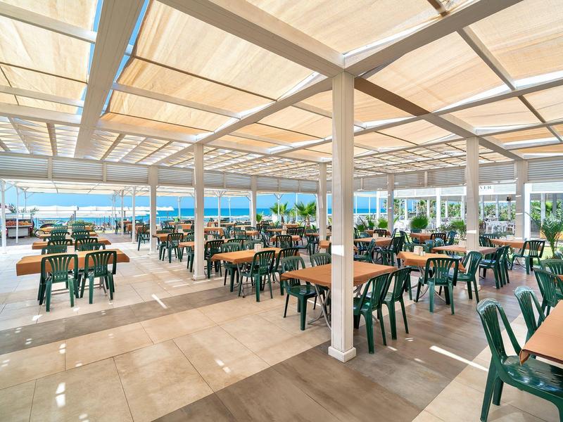 Amplia área exterior con muchas sillas verdes y mesas de madera bajo toldo blanco en hotel.