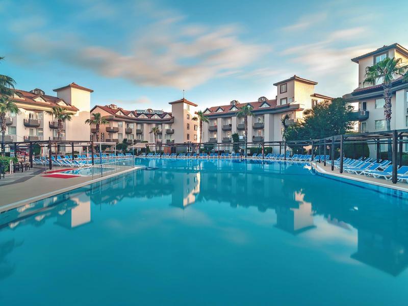 Amplia piscina en un hotel rodeado de edificios de varios pisos y vegetación bajo un cielo nublado.
