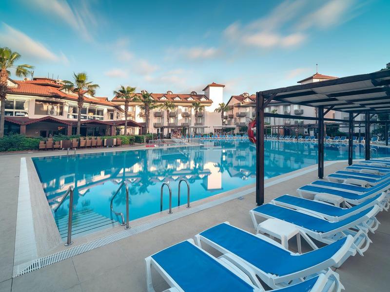 Una gran piscina de hotel con tumbonas azules y palmeras bajo un cielo azul.