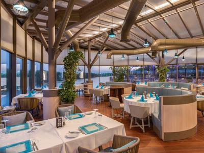 Modern eingerichtetes Restaurant mit Holzbalken, weißen Tischen und großen Fenstern bei Abendlicht.