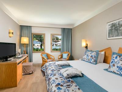 Hell eingerichtetes Hotelzimmer mit Doppelbett, TV, zwei Stühlen und Balkon mit Blick ins Grüne.