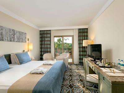 Helles Hotelzimmer mit Doppelbett, Schreibtisch, Stühlen und Fenster mit grünem Vorhang.