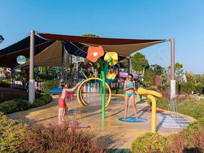 Kinder spielen an bunten Wasser-Spielgeräten unter Sonnensegeln bei klarem Himmel.