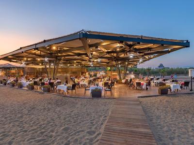 Offenes Strandrestaurant mit moderner Dachkonstruktion und Tischen im Sand bei Sonnenuntergang.