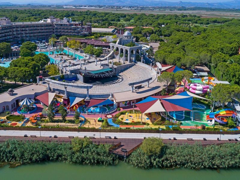 Parc aquatique avec piscines, toboggans et zones ombragées entouré d'arbres et d'un hôtel en arrière-plan