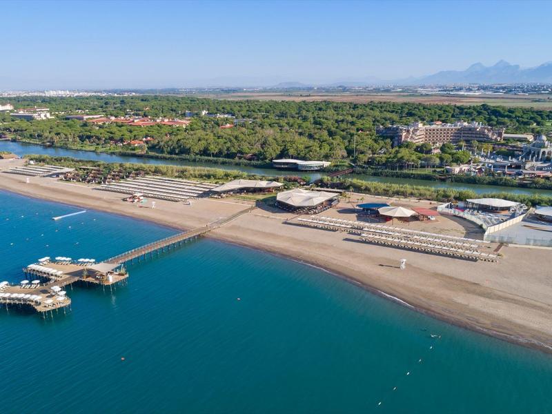 Vue aérienne d'une plage avec une jetée, une plage de sable et des hôtels en arrière-plan sous un ciel bleu clair.