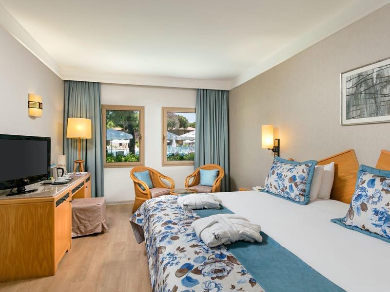 Chambre d'hôtel lumineuse avec lit double, TV, chaises et accès à une terrasse donnant sur une piscine.