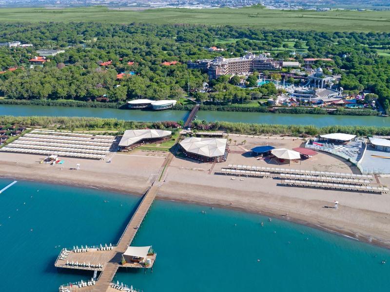 Plage avec jetée, rives sablonneuses, bâtiments entourés de verdure et montagnes en arrière-plan.