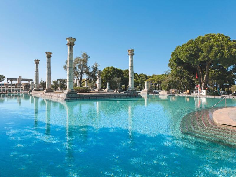 Grande piscine d'hôtel avec terrasse ensoleillée, colonnes antiques et arbres verts sous un ciel bleu.