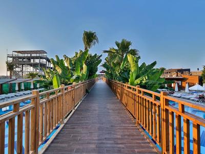 Szeroki drewniany pomost prowadzi do strefy basenowej z leżakami i palmami pod błękitnym niebem.