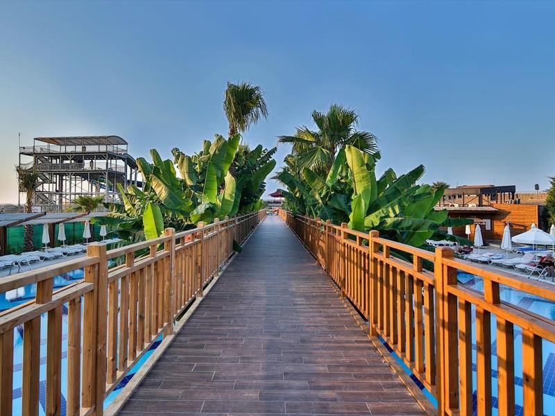 Široká dřevěná cesta vede k tropickému hotelu s bazény a palmami při západu slunce.