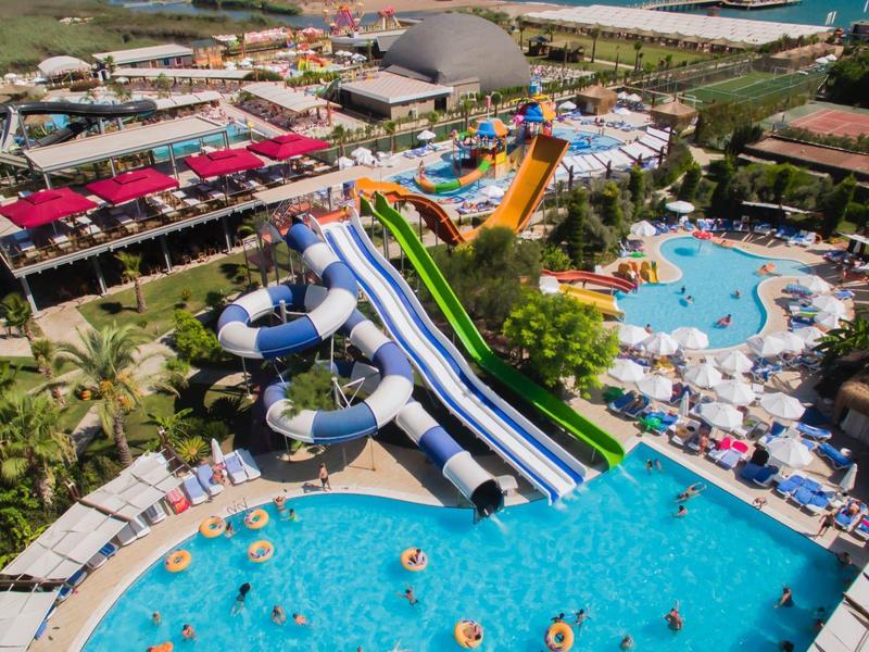 Pohled na velký vodní park s bazény, skluzavkami a lehátky pod jasnou oblohou.