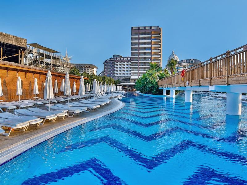 Velký venkovní bazén s modrou vodní plochou, lehátky a hotelovou budovou v pozadí.