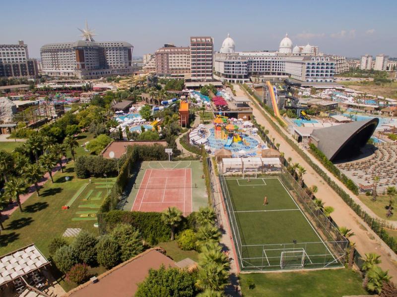 Letecký pohled na velké hotelové středisko s tenisovými kurty a upravenými zahradami za jasné oblohy.