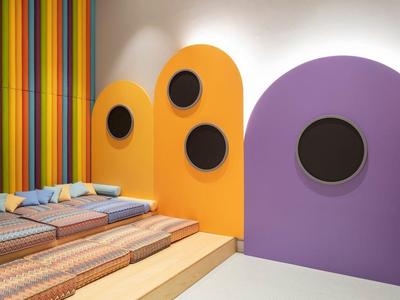 Area giochi moderna e colorata per bambini con sedute imbottite e TV a parete.