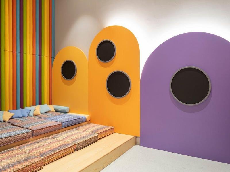 Area giochi moderna e colorata per bambini con sedute imbottite e TV a parete.