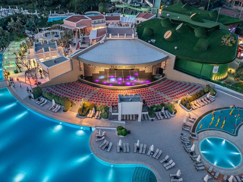 Resort notturno con piscina illuminata e anfiteatro, circondato da lettini e edifici.
