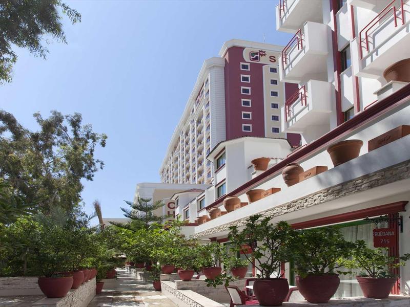 Moderní hotelová budova s venkovní terasou a květináči pod jasnou oblohou.