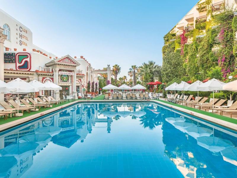 Hotelový bazén s lehátky a slunečníky mezi budovami pod modrou oblohou.