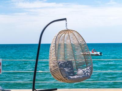Un fauteuil suspendu en osier sur une terrasse en bois avec vue sur la mer sous un ciel clair.