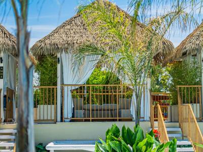 Bungalow de plage avec toit de chaume, palmiers et jardin vert sous un ciel bleu.