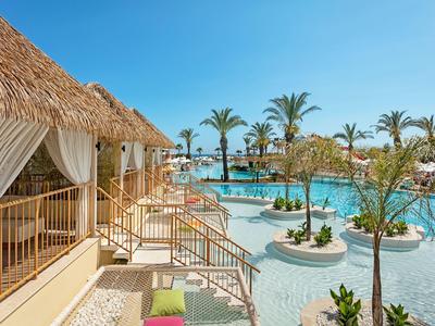 Espace piscine avec terrasse ensoleillée, palmiers et bungalows sous un ciel dégagé.