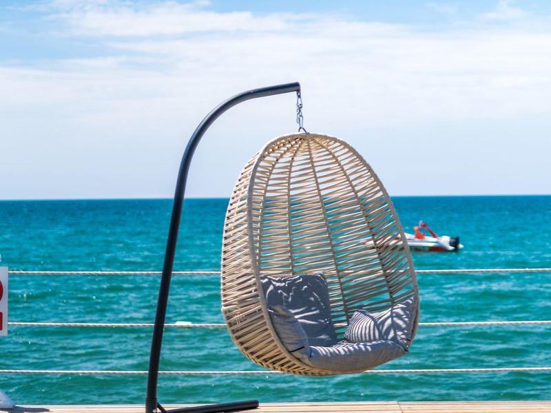 Un fauteuil suspendu en osier sur une terrasse en bois avec vue sur la mer sous un ciel clair.