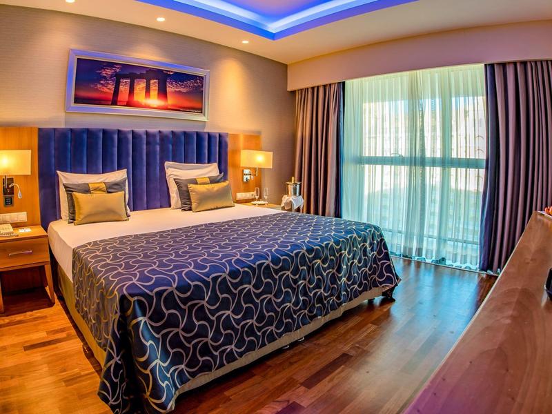 Lichte hotelkamer met tweepersoonsbed, blauwe accenten en groot raam met gordijnen.