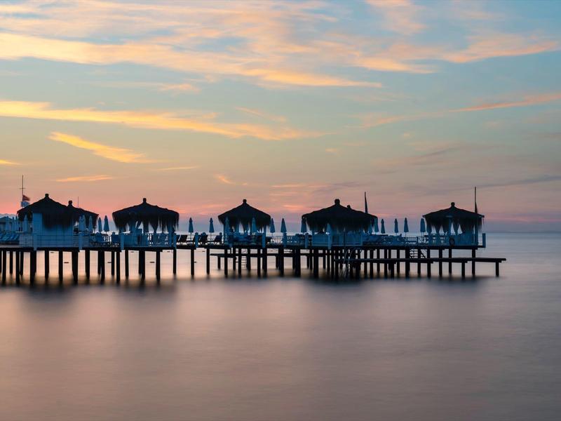 Zes kleine hutten op een lange pier boven kalm water bij zonsondergang.