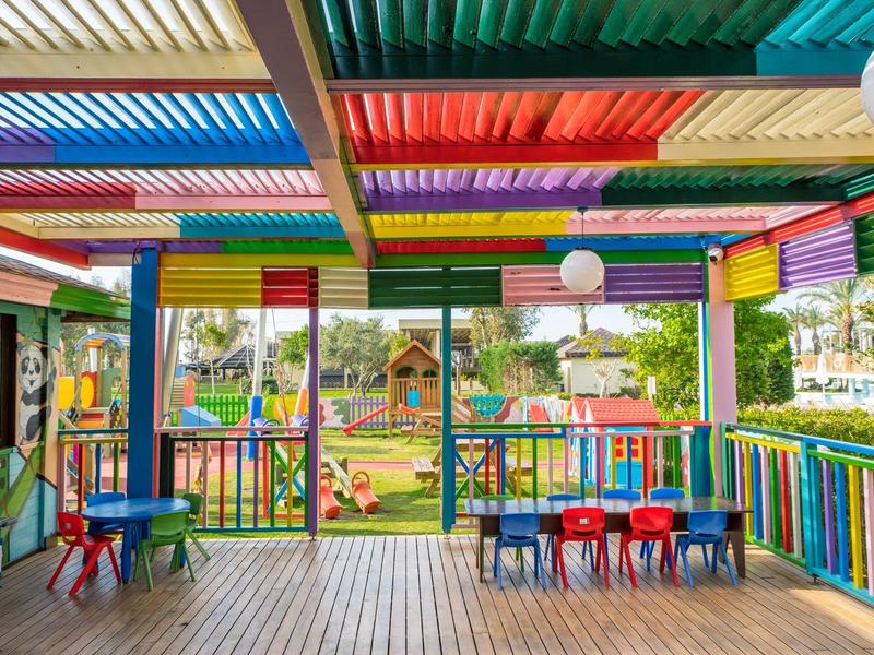 Kleurrijk overdekt buitengebied met kindertafels en uitzicht op een speeltuin.