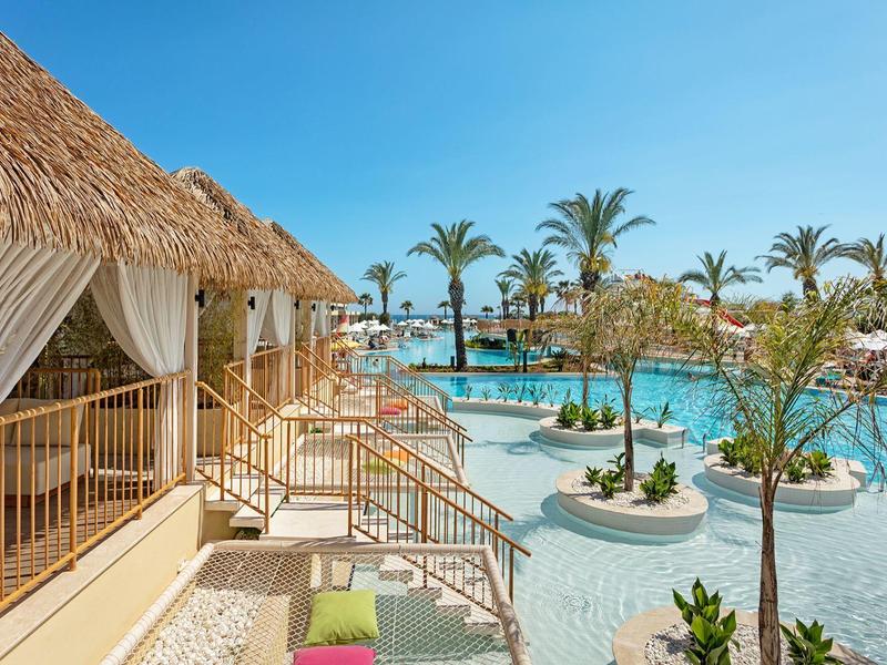 Espace piscine avec terrasse ensoleillée, palmiers et bungalows sous un ciel dégagé.