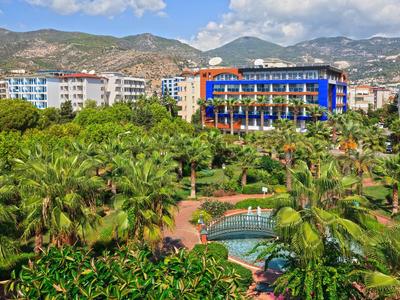 Grüner Park mit Palmen und Brunnen vor mehrstöckigen Wohngebäuden, Berge im Hintergrund.